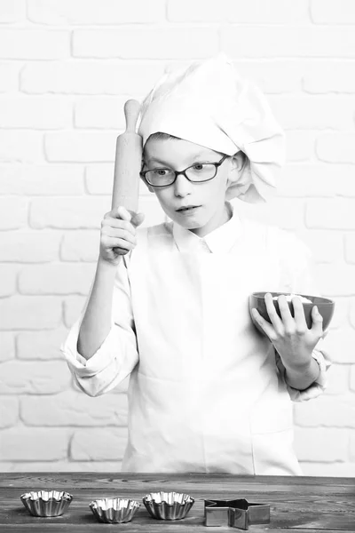 Genç çocuk küçük sevimli aşçı Şef beyaz üniforma ve kek için tablo kalıpları ile yakın duran ve oklava tuğla duvar arka plan üzerinde kırmızı bir kase tutarak gözlüklü ciddi yüz şapka — Stok fotoğraf