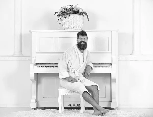 El hombre con barba en albornoz disfruta de la mañana mientras está sentado cerca del piano. El hombre alegre se sienta delante del instrumento musical de piano en el interior blanco sobre el fondo. Concepto de músico talentoso —  Fotos de Stock