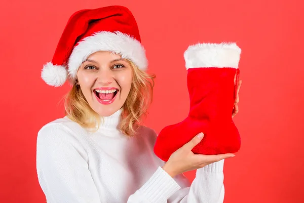 Girl cheerful face got gift in christmas sock. Check contents of christmas stocking. Traditional winter holiday. Woman in santa hat hold christmas gift red background. Christmas stocking concept