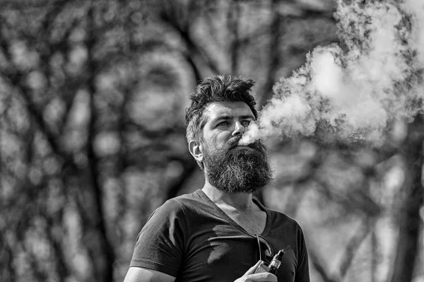 Conceito de cigarro eletrônico. Homem com barba longa e nuvens de fumaça parece relaxado. Homem com barba e bigode no rosto calmo, ramos no fundo, desfocado. Homem barbudo fuma vape no dia ensolarado — Fotografia de Stock