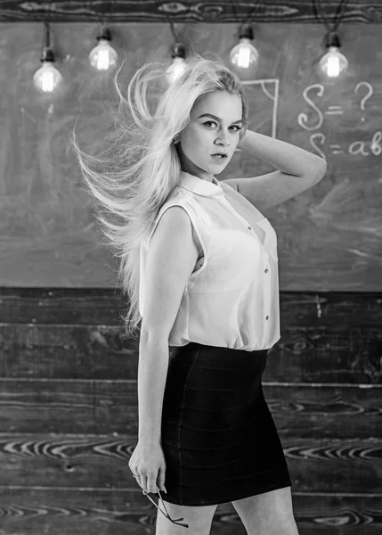 Sexy teacher concept. Teacher with waving long blonde hair looks sexy. Lady strict teacher on dreamy face stands in front of chalkboard. Woman with long hair in white blouse stands in classroom