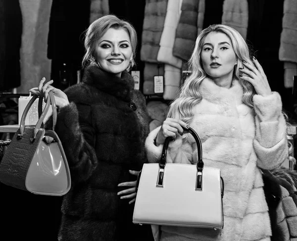 Girls with smiling faces in black and white fur coats hold purses in hands. Luxury style concept. Women with blond hair in fur coats with bags in fur shop. Women with makeup shopping in fashion store — Stock Photo, Image