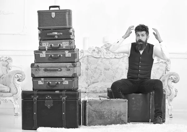 Macho elegant on surprised face sits shocked near pile of vintage suitcase. Luggage and relocation concept. Man, butler with beard and mustache delivers luggage, luxury white interior background