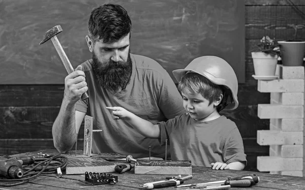 Boy, child busy in protective helmet learning to use hammer with dad. Father with beard teaching little son to use tools, hammering, chalkboard on background. Fatherhood concept