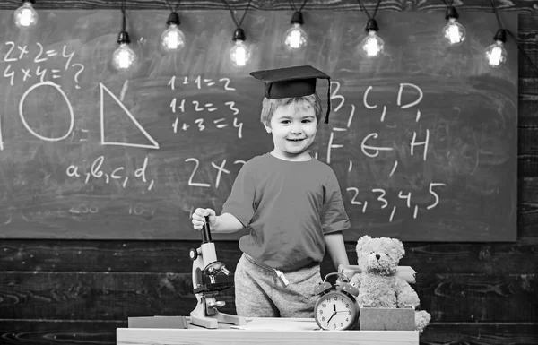 Kind, leerling op het glimlachen gezicht in de buurt van Microscoop. Eerste voormalige kochten studeren, onderwijs. Wonderkind concept. Kid jongen in graduate GLB in de buurt van Microscoop in klas, schoolbord op achtergrond — Stockfoto