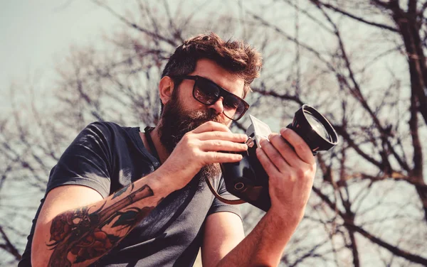 Reporter fotograf koncept. Mannen med skägg och mustasch bär solglasögon, sky på bakgrund. Guy skytte natur solig dag. Hipster på lugna ansikte innehar gamla gammaldags retro kamera — Stockfoto