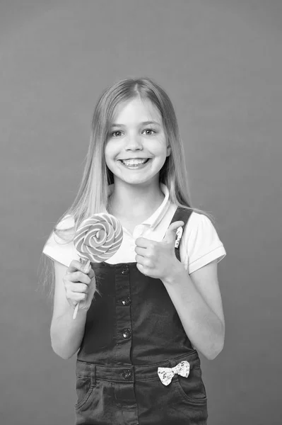 Mädchen essen bunte Lutscher. süße Kindheit Konzept. Kind mit langen Haaren mag Süßigkeiten. Mädchen mit lächelndem Gesicht hält riesige Bonbons in der Hand, violetten Hintergrund. Kind in rosa Overalls zeigt Daumen hoch — Stockfoto