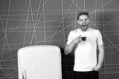 Uykulu esneyen adam sabah içkisini buzdolabının yanında tutuyor. Bachelor Hold fincanı pembe arka plandaki retro buzdolabında çay ya da kahve. Sabah çayı fikirleri ve tarifleri. Güne başlamak için sabah çayları