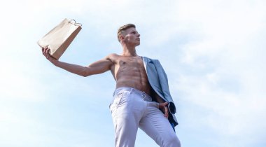 Adam çekici maço taşıma alışveriş çantası kahverengi kağıt yapılmış. Satış sezonu. Bu paketi al. Tezgâhtar kavramı. Adam kas atlet alışveriş çantası gökyüzü arka plan tutun. Sıcak satış ve indirim