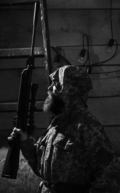 Avcı kavramı. Hunter, silahla acımasız hipster tüfek avcılık için hazırlayın. Maço gamekeepers evinde sıkı yüzünde. Sakallı adam başlıklı kamuflaj elbiseler, ahşap iç arka plan giyer