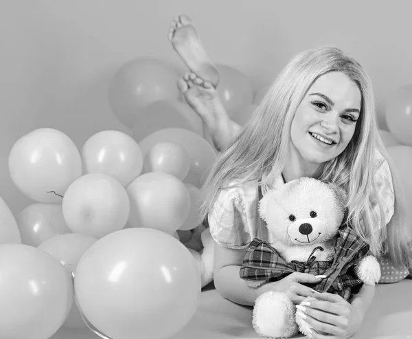 Birthday girl concept. Girl in pajama, domestic clothes lay near air balloons, pink background. Blonde on smiling face relaxing with teddy bear toy. Woman cute celebrate birthday with balloons