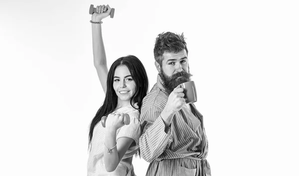 Casal apaixonado no pijama, posto de roupão isolado no fundo branco. Conceito alternativo matutino. Casal, família com rostos sonolentos, cheia de energia. Menina com haltere, homem com xícara de café — Fotografia de Stock