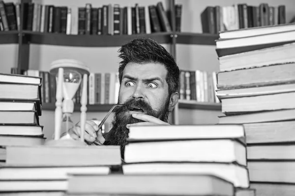 Man, scientist looks at hourglass. Man on serious and shocked face watching time is going over, bookshelves on background. Time flow concept. Teacher or student with beard studying in library