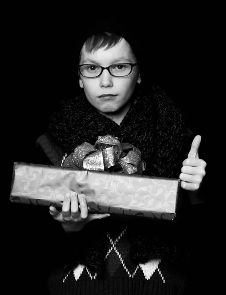 Petit garçon ou mignon nerd kid en lunettes, chapeau et foulard tricoté à la mode sur fond noir tient boîte cadeau rouge avec arc doré et pouce u — Photo