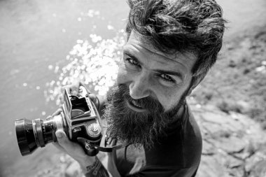 Sakal ve bıyık tutar kamera, güneşli, su yüzeyine arka plan açık olan adam. Fotoğrafçı kavramı. Hipster gülümseyen yüzü üzerinde eski moda kamera tutar. Nehir kenarında doğa çekim adam