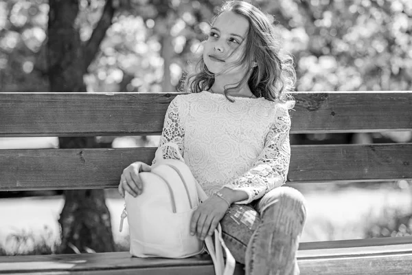 Girl on dreamy face sits on bench, sakura tree on background, defocused. Spring walks concept. Cute child with backpack enjoy sunny spring day. Girl relaxing while walk in park near cherry blossom — Stock Photo, Image