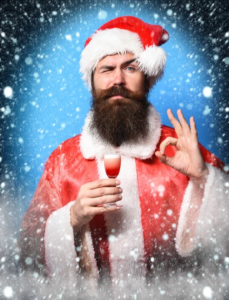 Guapo barbudo hombre de Santa Claus con barba larga en la cara seria sosteniendo vaso de trago alcohólico en navidad roja o suéter de Navidad y sombrero de año nuevo que muestra ok en fondo de estudio azul —  Fotos de Stock