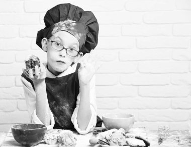 Genç çocuk sevimli aşçı Şef kırmızı üniforma ve renkli kaseler, lezzetli çerezler, oklava ile masada oturan ve çikolatalı kek beyaz tuğla duvar adam üzerinde tutarak gözlüklü lekeli yüz şapka