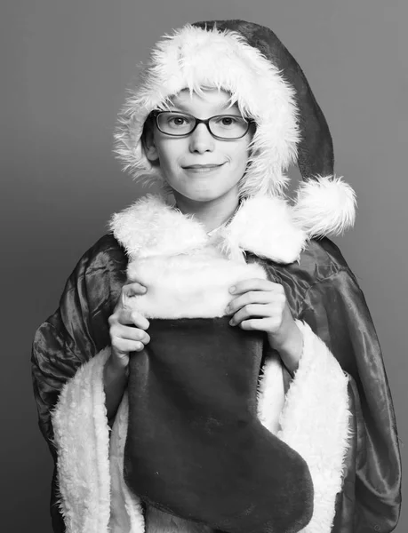 Joven lindo niño de Santa Claus con gafas en suéter rojo y sombrero de año nuevo sostiene la Navidad decorativa o medias de Navidad o bota en el fondo del estudio —  Fotos de Stock