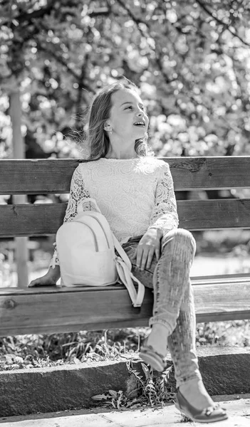 Dziewczyna na uśmiechniętą twarz siedzi na ławce, drzewa sakura na rozmytym tłem. Słodkie dziecko z plecaka cieszyć się słoneczny wiosenny dzień. Relaks podczas spaceru w parku, w pobliżu wiśniowy kwiat dziewczyna. Wiosna idzie koncepcja — Zdjęcie stockowe