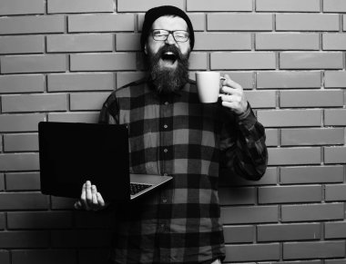 Sakallı, beyaz tenli hippi elinde bardakla dizüstü bilgisayar tutuyor.