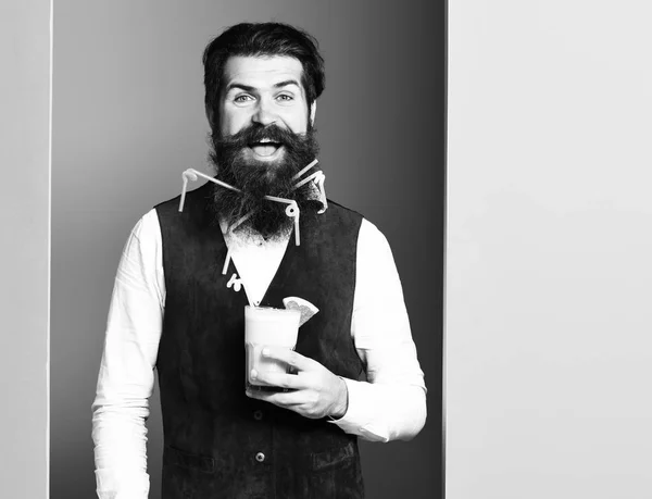 handsome bearded man with long beard and mustache has stylish hair on smiling face holding glass of alcoholic beverage in vintage suede leather waistcoat on colorful studio background