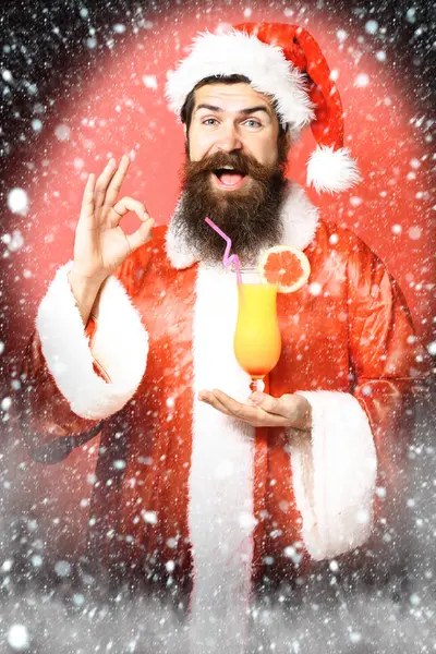 Bonito barbudo santa claus homem com longa barba no rosto feliz segurando vidro de coquetel não alcoólico no Natal ou xmas suéter e chapéu de ano novo mostrando ok no fundo estúdio vermelho — Fotografia de Stock