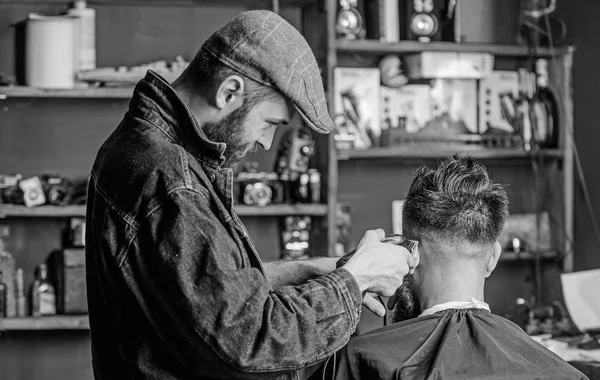 Barber with clipper trimming hair of client, rear view. Barber with hair clipper works on short haircut on nape, barbershop background. Hipster client getting short haircut. Working process concept