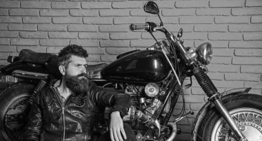 Sakallı, motorcu motorlu bisiklet garaj, tuğla duvar arka plan yakınındaki deri ceketli adam. Bisikletçileri yaşam tarzı kavramı. Yenilikçi, vahşi Motorcu deri ceketli dalgın yüzünde oturuyor, motosiklet üzerinde eğilir