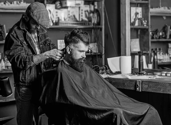 Barber with hair clipper works on haircut of bearded guy, retro barbershop  background. Hipster hairstyle concept. Hipster client getting haircut.  Barber with clipper trimming hair on nape of client - Stock Image -