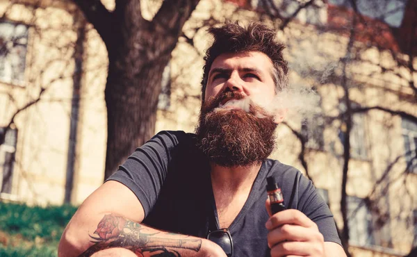 Hipster con el pelo alborotado y gris en la cara de ensueño relajado con humo blanco que vuela por la boca. Concepto de aspiración y hábitos. Hombre con barba y bigote fumar, fondo urbano —  Fotos de Stock