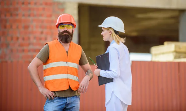 Woman inspector and bearded brutal builder discuss construction progress. Construction project inspecting. Safety inspector concept. Construction site safety inspection. Discuss progress project