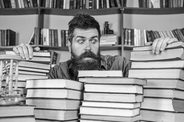 Profesor o estudiante con barba se sienta a la mesa con libros, desenfocado. Hombre en la cara conmocionada entre montones de libros, mientras estudiaba en la biblioteca, estanterías en el fondo. Concepto bibliófilo —  Fotos de Stock