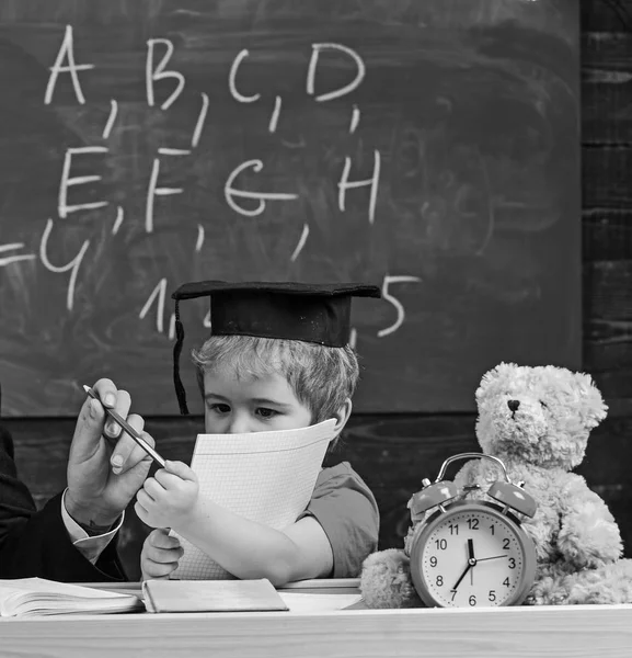 Grundutbildning koncept. Elev i mortarboard, svarta tavlan på bakgrund. Kid studier med häfte, manlig hand ger pennan. Pojken vill skriva eller rita med penna. Barn lära sig att skriva — Stockfoto