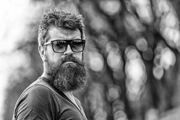 Barbearia e conceito de estilo. Homem com barba e bigode no rosto estrito, fundo da natureza, desfocado. O homem barbudo usa óculos de sol modernos. Hipster com barba parece confiante enquanto está de pé ao ar livre — Fotografia de Stock