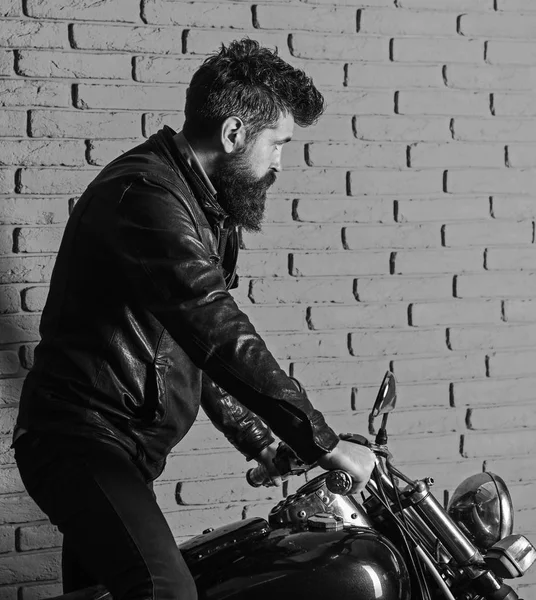 Homem com barba, motociclista em jaqueta de couro perto de moto motor na garagem, fundo da parede de tijolo. Início do conceito de viagem. Hipster, motociclista brutal no rosto sério em jaqueta de couro fica na motocicleta — Fotografia de Stock