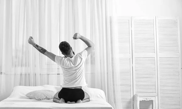 Guy stretching arms, full of energy in morning. Macho with beard stretching, relaxing after nap, rest. Man in shirt sits on bed, white curtains on background, rear view. Good morning concept