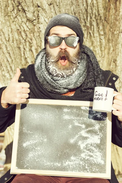 Stilig lycklig man, hipster, med skägg och mustasch i svarta solglasögon täckt med vit frost håller tom svarta tavlan, tummen upp och kaffekopp vinterdag på naturlig bakgrund, kopia spac — Stockfoto