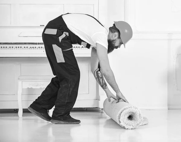 Relocating concept. Man with beard, worker in overalls and helmet rolling carpet, white background. Loader wrappes carpet into roll. Courier delivers furniture in case of move out, relocation