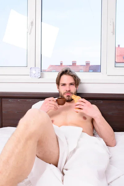 Чоловік бородатий красивий хлопець їсть сніданок у ліжку. Хлопець має каву і тримає круасан, лежачи в спальні або готельному номері. Чоловік їсть круасан і п'є каву. Приємний початок дня — стокове фото