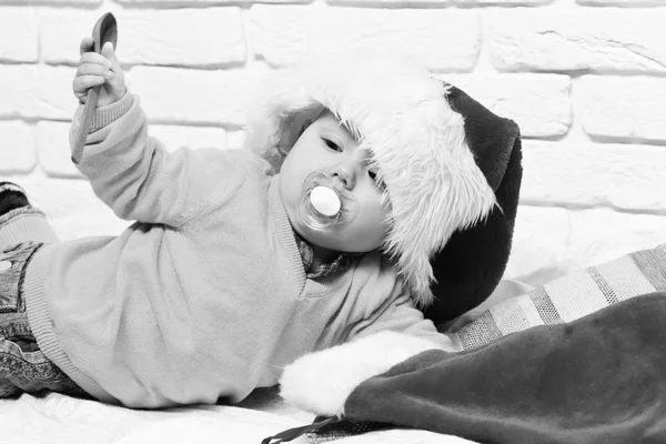 Mały chłopiec z uroczą ciekawą twarzą w żółtym swetrze leżącym na brzuchu z manekinem w kapeluszu noworocznym i Boże Narodzenie lub świąteczne pończochy lub buty na białym tle ściany z cegły — Zdjęcie stockowe