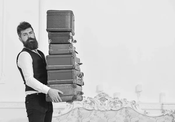 Άνδρας με μούσι και μουστάκι που φοράει το κλασικό κοστούμι παραδίδει αποσκευές, πολυτελή λευκό εσωτερικό υπόβαθρο. Macho, κομψό porter μεταφέρει βαρύ σωρό vintage βαλίτσες. Υπηρεσία μπάτλερ και έννοια — Φωτογραφία Αρχείου