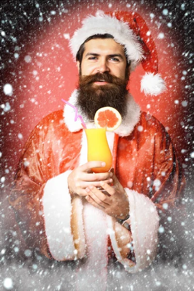 Όμορφος γενειοφόρος άνδρας Βασίλη με μακριά γενειάδα για χαρούμενο πρόσωπο κρατά ποτήρι μη αλκοολούχα κοκτέιλ στο Χριστούγεννα ή Χριστούγεννα πουλόβερ και το νέο έτος με καπέλα σε φόντο κόκκινο στούντιο — Φωτογραφία Αρχείου