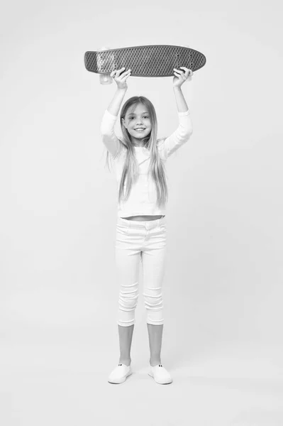 Menina segurando skate violeta acima de sua cabeça. Campeão na corrida de rua, conceito de infância feliz. Miúdo de salto branco, jeans e ténis isolados no fundo rosa. Criança com um rosto muito sorridente — Fotografia de Stock