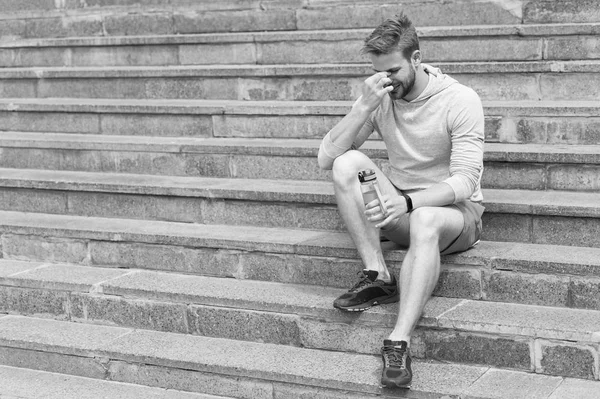 Tired after training on fresh air. Stay hydrated and healthy. Macho with water bottle sit on steps, copy space. Thirst. Strong man with plastic flask relax on stairs. Sport and fitness