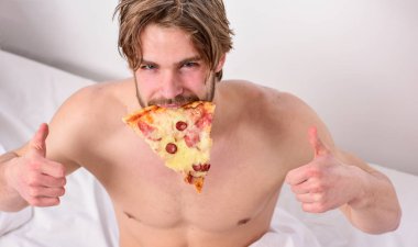 Adam, kahvaltıda pizza seviyor. Diyet hakkında kimin umurunda. Yakışıklı adam bir parça pizza elinde tutar ve bu yemek. Pizza yatakta.