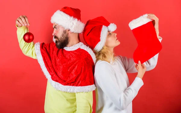 Kvinna och skäggig man i santa hatt väntar jul röd bakgrund. Par christmas santa dräkt håll strumpa och prydnad bollen. Julstrumpa tradition. Christmas celebration koncept — Stockfoto