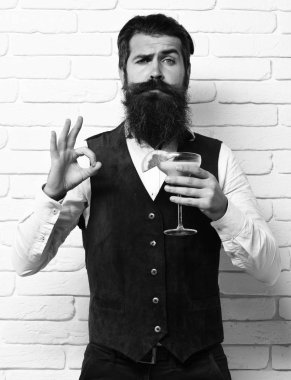 Uzun sakallı ve bıyıklı yakışıklı bir adamın ciddi yüzünde şık saçları var. Beyaz tuğla duvarlı stüdyo arka planında klasik süet deri bir yelek içinde alkollü içecek bardağı tutuyor.