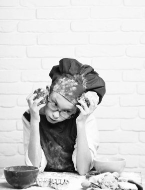 Genç çocuk sevimli aşçı Şef kırmızı üniforma ve renkli kaseler, lezzetli çerezler, oklava ile masada oturan ve çikolatalı kek beyaz tuğla duvar adam üzerinde tutarak gözlüklü lekeli yüz şapka