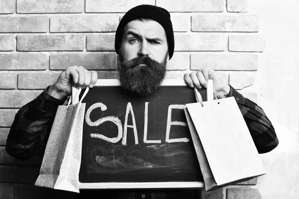 Bearded hipster holding shopping packages and board with Sale inscription — Stock Photo, Image